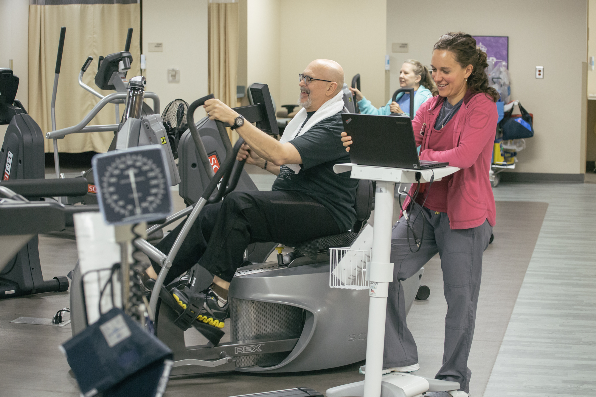 An older man on a stationary bicycle, coached by a physical therapist